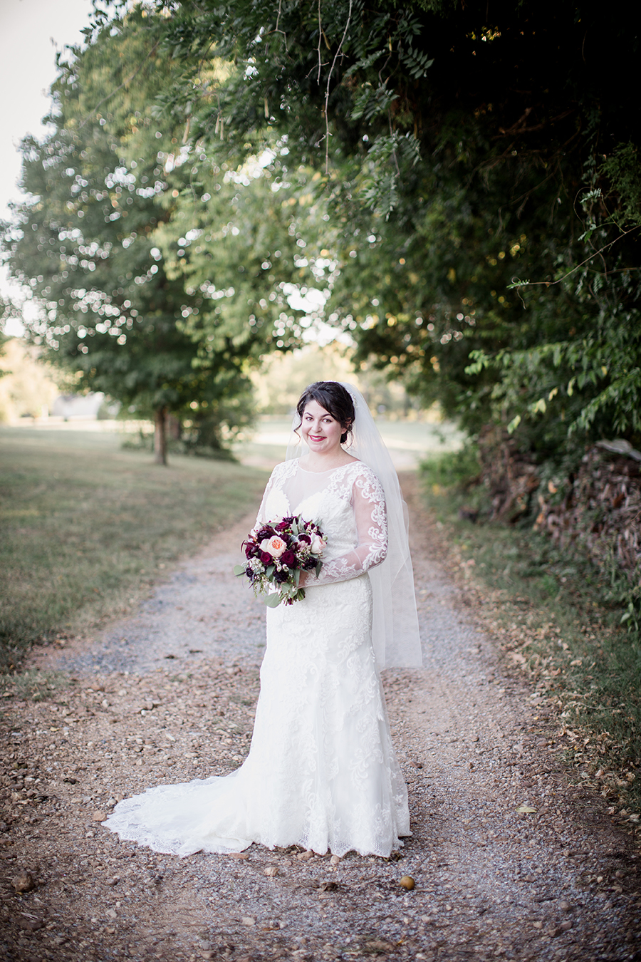 Looking at camera smiling holding her bouquet on gravel drive at this bridal session at The Barn at High Point Farms by Knoxville Wedding Photographer, Amanda May Photos.