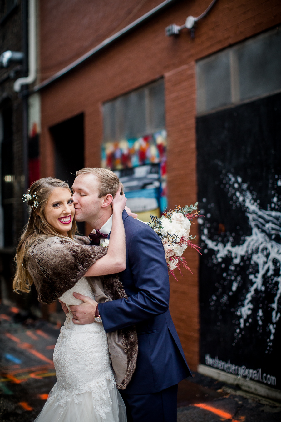 He kisses her cheek in the art alley during the bride and groom romantic portraits at this winter wedding at Knoxville Wedding Venue, Jackson Terminal, by Knoxville Wedding Photographer, Amanda May Photos.