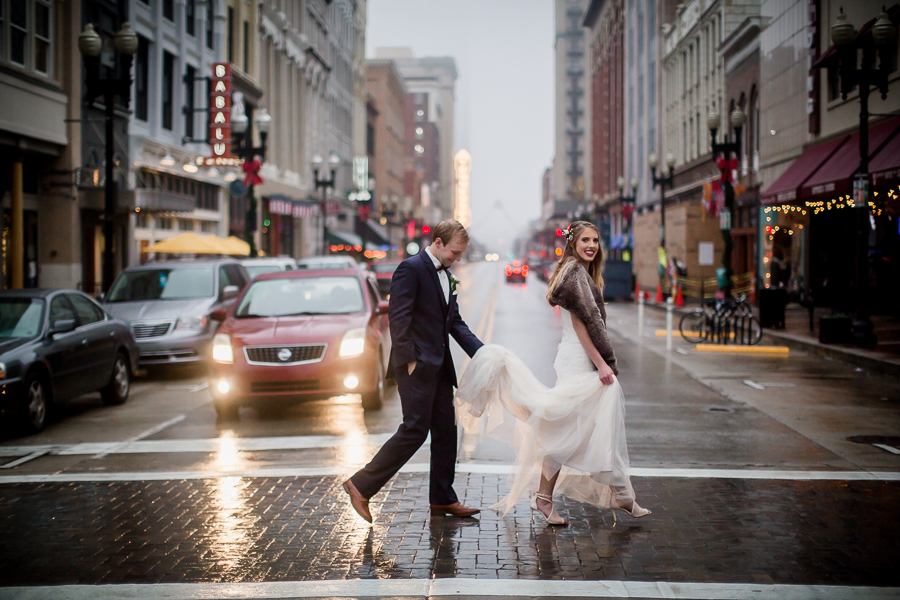Groom carries the bride's dress crossing the street during the bride and groom romantic portraits at this winter wedding at Knoxville Wedding Venue, Jackson Terminal, by Knoxville Wedding Photographer, Amanda May Photos.